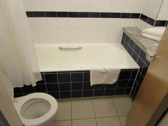 a typical bathroom in the Royal National Hotel with white tiles on the walls and floor. The bathroom is compact, but it's equipped with all the essentials, including a toilet, sink, and shower/tub combo. The sink is mounted on a small vanity with a mirror above it, and there's a towel rack nearby for convenience.