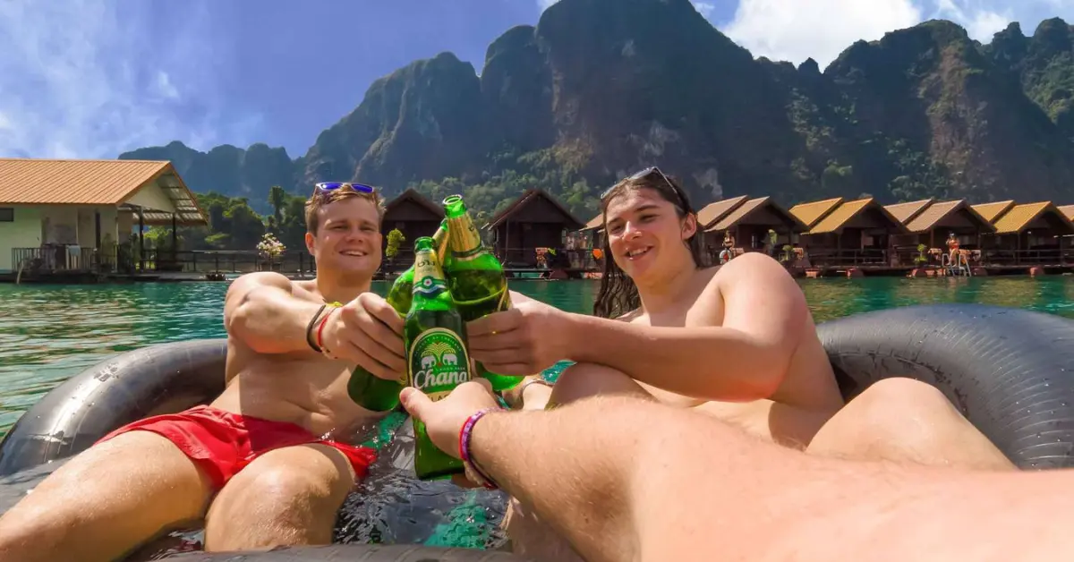 Two young travellers cheerfully clink Chang beer bottles while floating on a rubber tube in a serene lake with floating huts and limestone cliffs in the background.