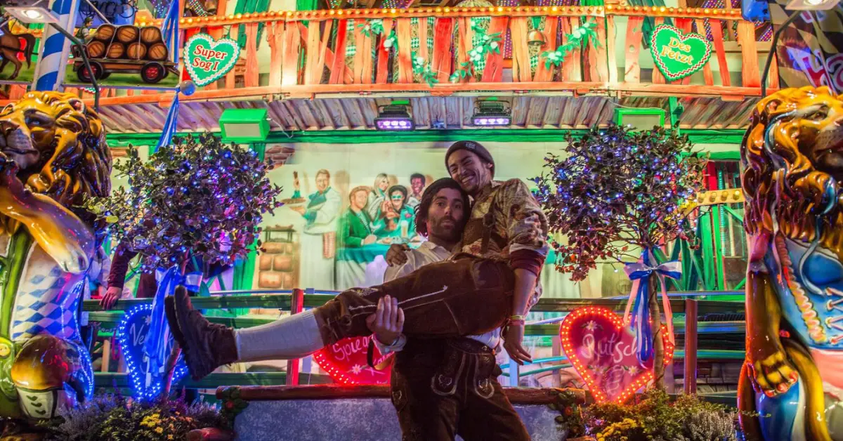 Two jubilant travellers enjoying a festival atmosphere, with one carrying the other in a playful pose in front of a vibrant carousel.