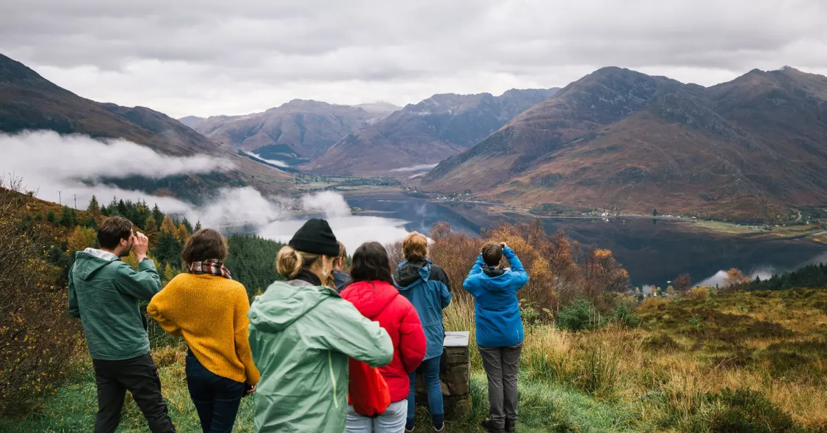 A group of young adults in colourful outdoor gear admiring a misty mountainous landscape with a lake below