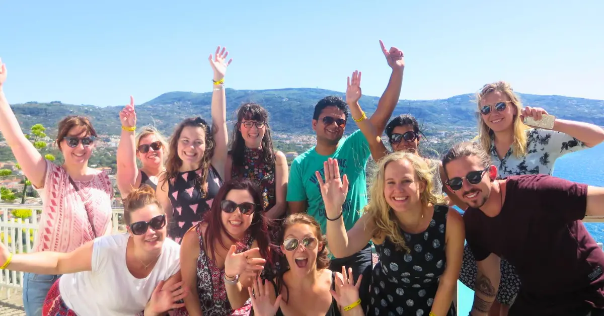 A group of cheerful young adults posing for a photo with their arms raised in joy, overlooking a scenic hillside.