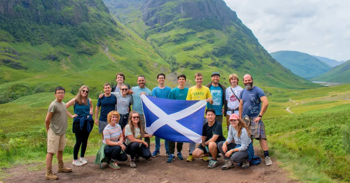 A diverse group of travellers holding a Scottish flag with lush green hills in the background on a MacBackpackers affordable tour of Scotland.