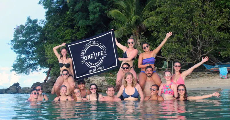 Joyful travellers holding a One Life Adventures banner while standing in tropical waters off an Asian beach.