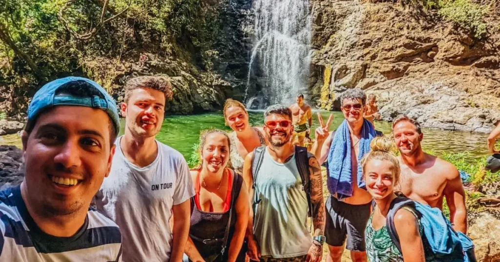A group of young adults on a TruTravels tour, smiling and posing for a selfie in front of a picturesque waterfall, embodying the adventurous spirit of youth-oriented travel experiences