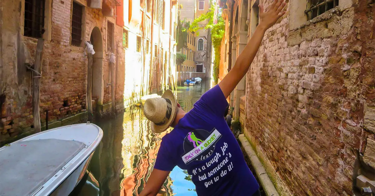 A tour guide from Italy on a Budget Tours is joyfully reaching up in a narrow, sunlit Venetian canal.