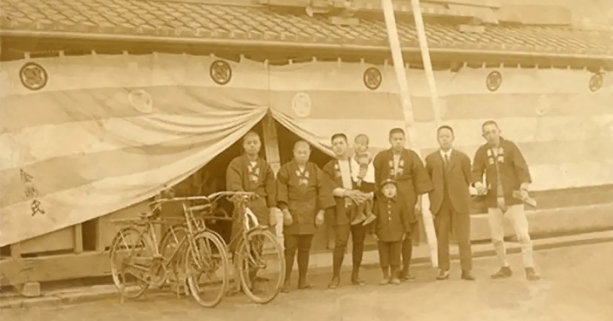 Sepia-toned historic photo of Japanese individuals and children in front of a building, with bicycles, under the banner of Kongo Gumi, showcasing an interesting fact about Japan's oldest company.