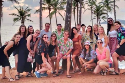 A cheerful group of travellers from One Life Adventures posing together on a tropical beach at sunset, with palm trees and the ocean in the background.