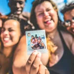 A group of six smiling friends poses closely together at a pool or beach, holding a polaroid photo of themselves. The background features bright blue skies and water, creating a cheerful atmosphere. The group showcases friendship on their Contiki Reunion Trips and joy, with vibrant swimwear and a lively vibe.