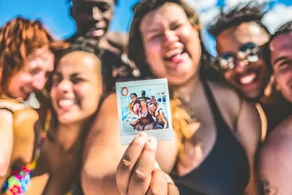 A group of six smiling friends poses closely together at a pool or beach, holding a polaroid photo of themselves. The background features bright blue skies and water, creating a cheerful atmosphere. The group showcases friendship on their Contiki Reunion Trips and joy, with vibrant swimwear and a lively vibe.