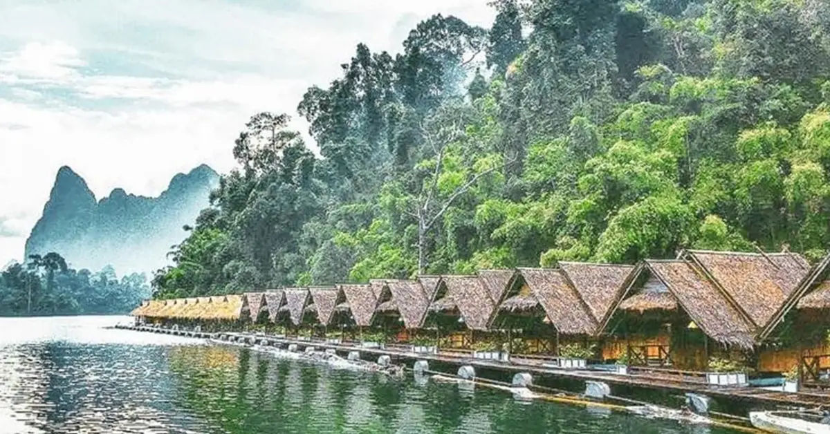 A row of thatched-roof huts on stilts extends over a calm body of water, with lush green forest and mist-covered mountains in the background. The serene scene is bathed in soft, filtered light from a partly cloudy sky.