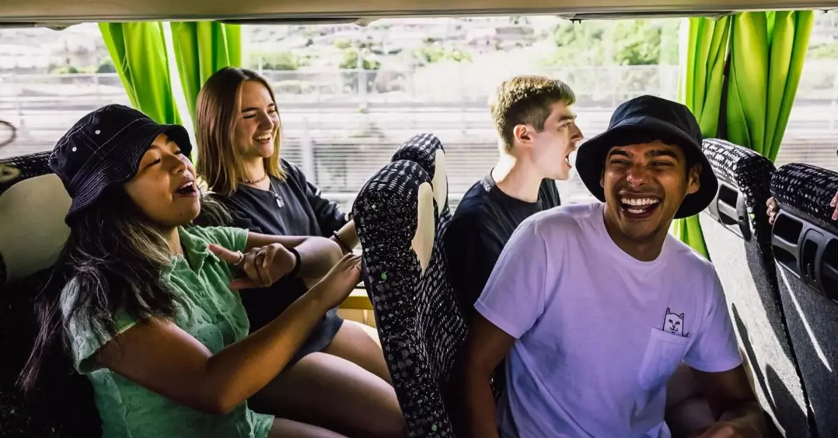 A group of four young friends enjoying a bus ride. On the left, a person in a black bucket hat and green shirt is animatedly talking or singing. Next to them, a person with long hair in a black shirt is smiling. Across the aisle, a person with short hair in a white shirt is laughing or talking. On the far right, another person in a black bucket hat and white shirt is laughing heartily. The bus features green curtains and patterned seat covers, with a blurred view of greenery and a cityscape outside the window, indicating the bus is in motion. The atmosphere is cheerful and lively.