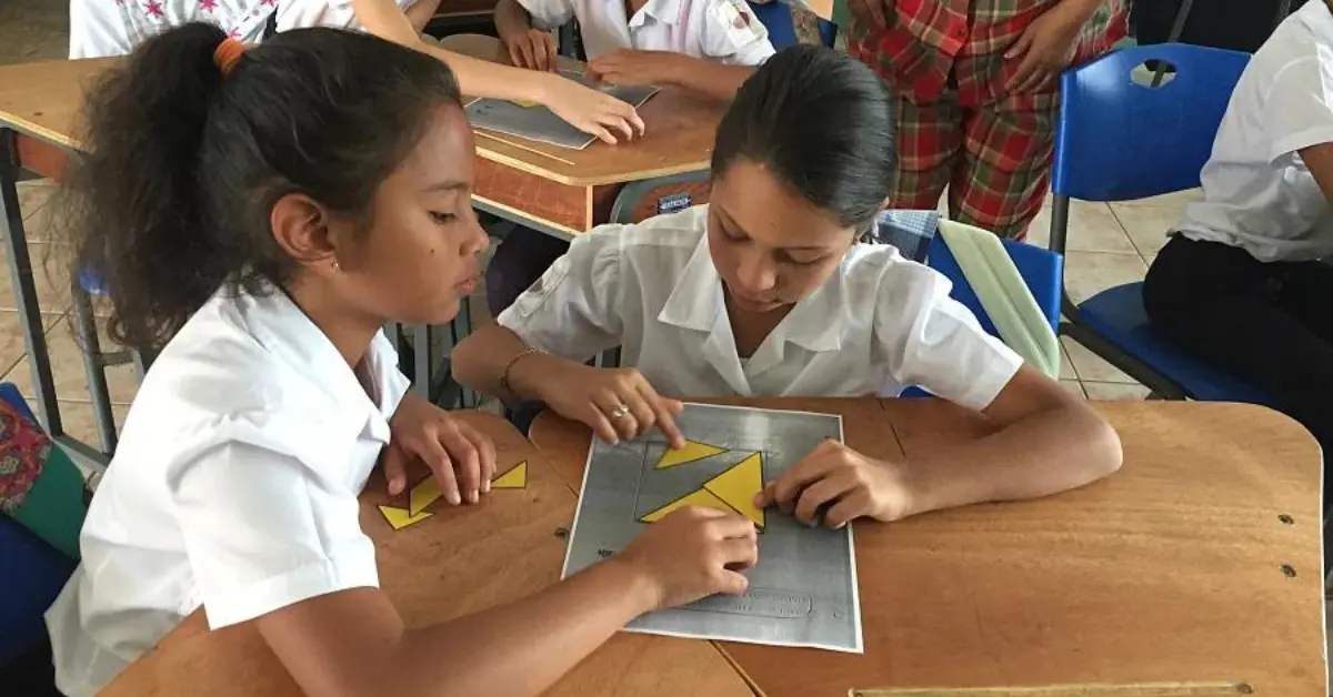 Two schoolgirls in white uniforms working together on a tangram puzzle at a wooden desk in a classroom. Other students are visible in the background, also engaged in activities. The scene depicts a collaborative and educational environment.