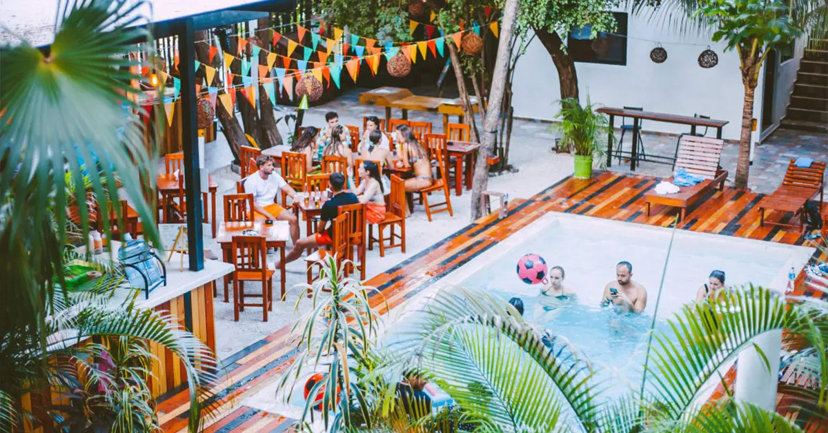 Vibrant outdoor gathering at a G Adventures Tours Roamies event, featuring a lively dining area under colorful flags and a nearby pool surrounded by lush greenery, capturing the essence of adventure and social connection