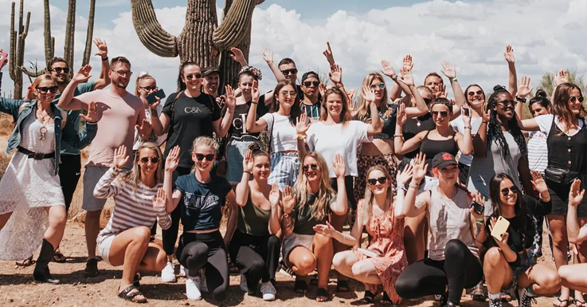 Group of happy travelers posing in front of a large cactus during a sunny desert adventure with Topdeck Tours. The diverse group, dressed in casual summer attire, smiles and waves at the camera, capturing the fun and excitement of their outdoor exploration. Blue skies and a scenic desert backdrop highlight the vibrant and joyful atmosphere of this unforgettable travel experience.