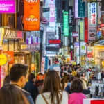 Photograph of a bustling street in South Korea, filled with people walking and shopping. The street is lined with numerous shops and stalls, displaying colourful signs and advertisements in Korean. Neon lights and vibrant shopfronts create a lively atmosphere. The crowd includes both locals and tourists, contributing to the energetic vibe of the market area.