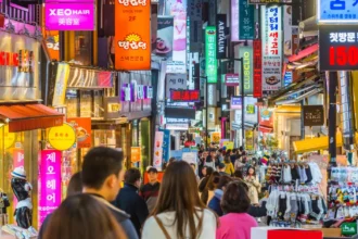 Photograph of a bustling street in South Korea, filled with people walking and shopping. The street is lined with numerous shops and stalls, displaying colourful signs and advertisements in Korean. Neon lights and vibrant shopfronts create a lively atmosphere. The crowd includes both locals and tourists, contributing to the energetic vibe of the market area.