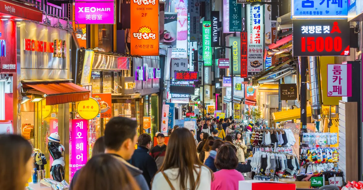 Photograph of a bustling street in South Korea, filled with people walking and shopping. The street is lined with numerous shops and stalls, displaying colourful signs and advertisements in Korean. Neon lights and vibrant shopfronts create a lively atmosphere. The crowd includes both locals and tourists, contributing to the energetic vibe of the market area.