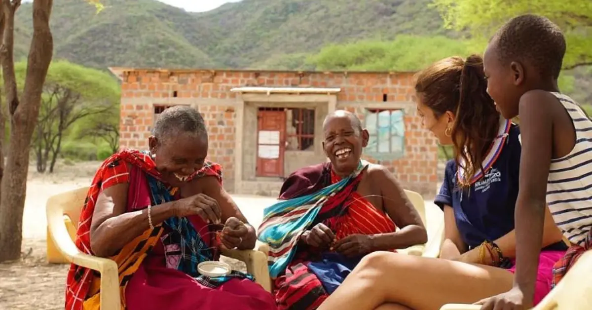 Two elderly women in colourful traditional clothing are sitting outdoors, smiling and beading jewellery. They are joined by a young woman and a child, both of whom are also smiling. In the background, there is a brick building and lush green hills. The scene is set in a rural area with trees and mountains.