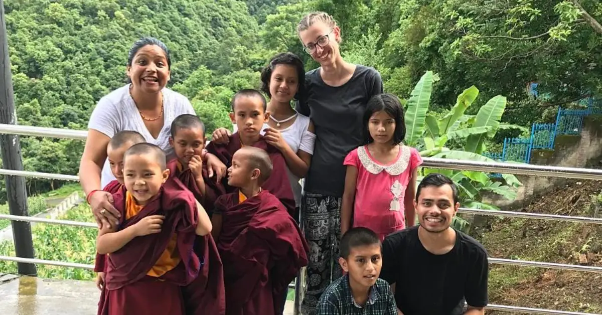 A group of children and adults posing for a photo on a balcony with lush green trees in the background. The children, some of whom are wearing maroon robes, are standing and sitting alongside the adults, who are smiling. The setting appears to be a rural or mountainous area, with a serene and natural environment.