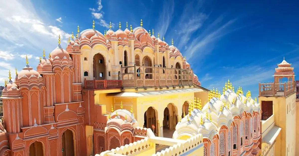 A vibrant view of the Hawa Mahal, also known as the Palace of Winds, in Jaipur, India. The palace features intricate pink and orange sandstone architecture with numerous small windows and ornate balconies. The sky is clear and blue, adding to the striking beauty of the historic structure.