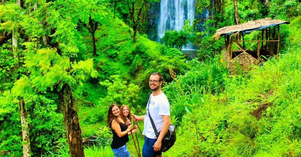 Three people smiling and posing for a photo in a lush green forest with a waterfall in the background. They are standing on a grassy path, with one person holding a walking stick. To the right, there is a small thatched hut. The scene is vibrant and full of greenery, suggesting a tropical or subtropical location.