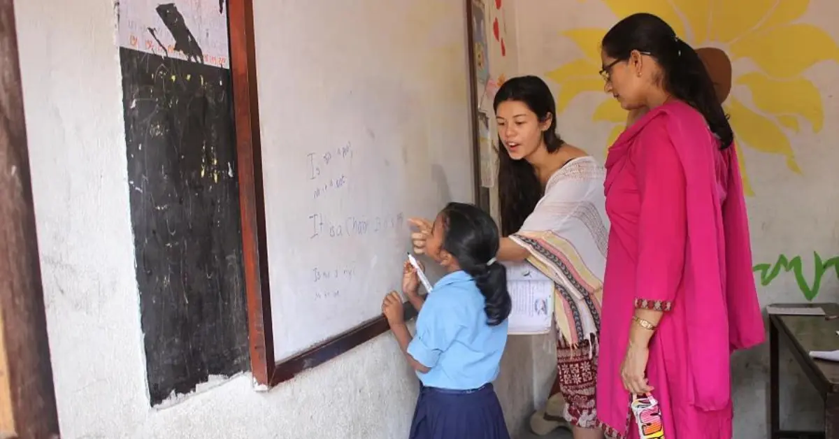 Many young adults seek volunteer work overseas to teach English. The image shows a young woman assisting a child at a whiteboard, with another adult observing. The child is writing in English on the board, and the setting appears to be a classroom with educational posters and a colourful mural on the wall.