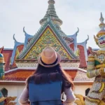 A tourist wearing a hat stands in front of a traditional Thai temple, flanked by two colourful guardian statues.