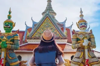 A tourist wearing a hat stands in front of a traditional Thai temple, flanked by two colourful guardian statues.