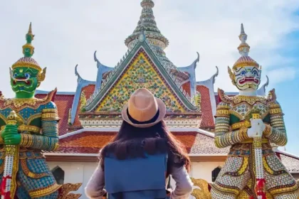 A tourist wearing a hat stands in front of a traditional Thai temple, flanked by two colourful guardian statues.