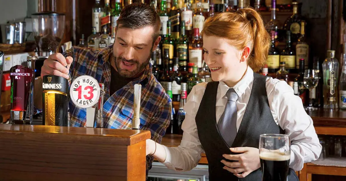 Young adult working in an Irish pub on a working holiday, learning to pour drinks from a friendly bartender behind a well-stocked bar