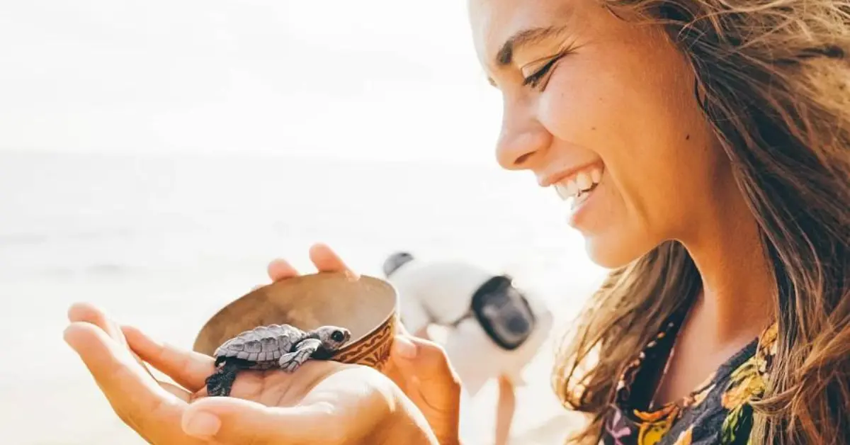 A woman smiling and holding a small sea turtle in her hands on a beach. She is using a bowl to support the turtle. In the background, another person is crouching down, possibly taking a photo or observing the scene. The beach and ocean are visible, suggesting a coastal environment. The image conveys a sense of joy and connection with nature.