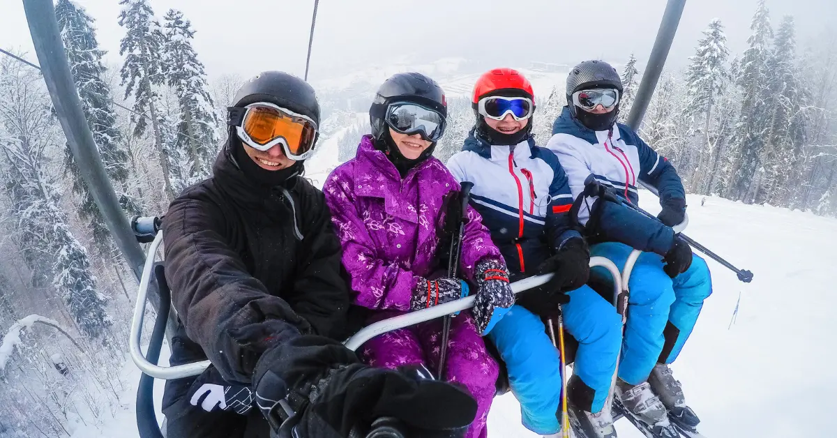 Four people on a ski lift, dressed in winter sports gear, smiling and enjoying a snowy day in Canada. They are on a working holiday, taking a break to enjoy the slopes. The background features snow-covered trees and a mountainous landscape.