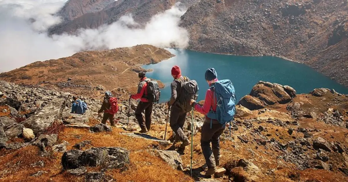 A group of hikers descending a rocky mountain trail towards a serene blue lake surrounded by rugged terrain. The hikers are equipped with backpacks and walking sticks, dressed in warm clothing and hats. The background features misty clouds partially covering the mountain peaks, creating a breathtaking and adventurous landscape.