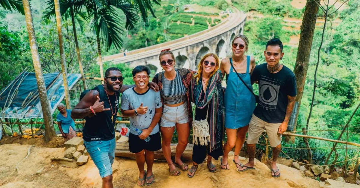 Group of six travellers posing together in front of the iconic Nine Arches Bridge in a lush, green landscape. The cheerful group, dressed in casual summer attire, enjoys the scenic view of the historic stone bridge and terraced fields, highlighting the joy of exploration and adventure.