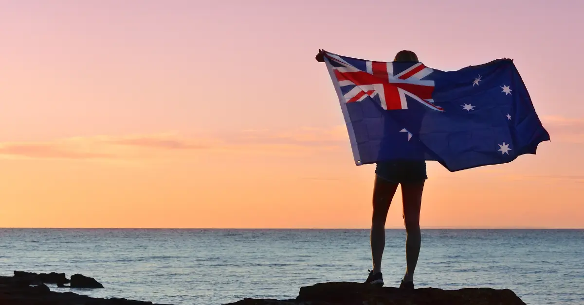 A person standing on a rocky shoreline, holding an Australian flag against a stunning sunset. The sky is painted in shades of pink, orange, and yellow, with the calm sea reflecting the warm colours. The scene evokes a sense of patriotism and tranquillity.