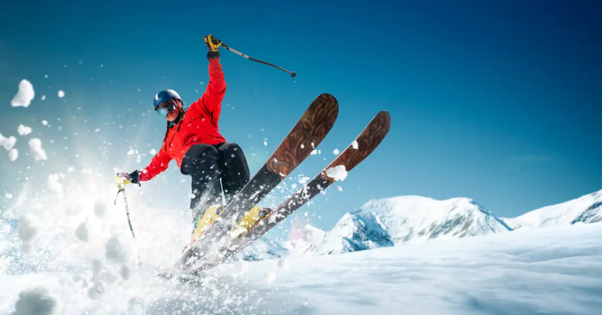 A skier in a red jacket and black helmet is captured mid-air while skiing down a snowy slope, with snow spraying around them. The background features a clear blue sky and snow-covered mountains.