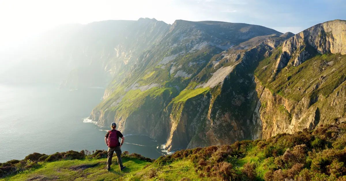 A hiker stands on a grassy cliff edge, overlooking a stunning coastal landscape with towering cliffs and the sea below, bathed in the soft light of the setting sun.