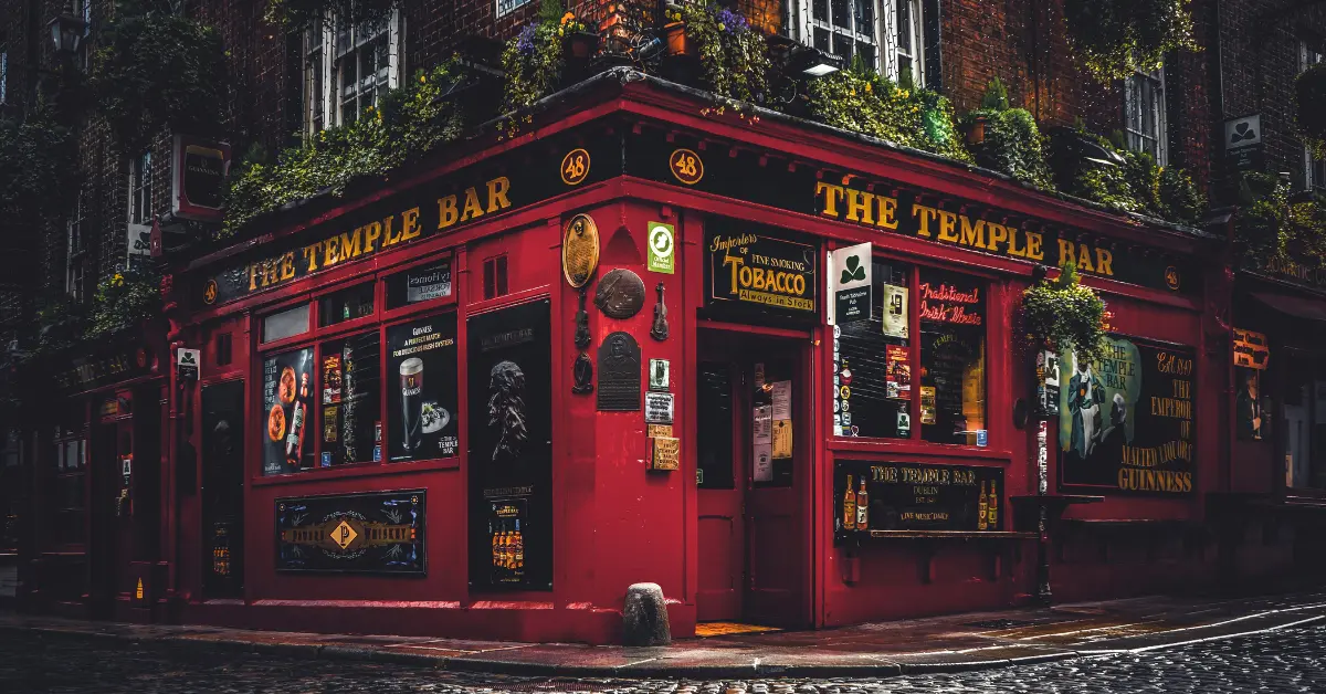 A vibrant red pub named "The Temple Bar" located on a cobblestone street corner. The exterior is adorned with various signs, posters, and hanging plants. The pub has large windows displaying advertisements for drinks and traditional Irish music. The scene captures the charm and character of this iconic establishment.