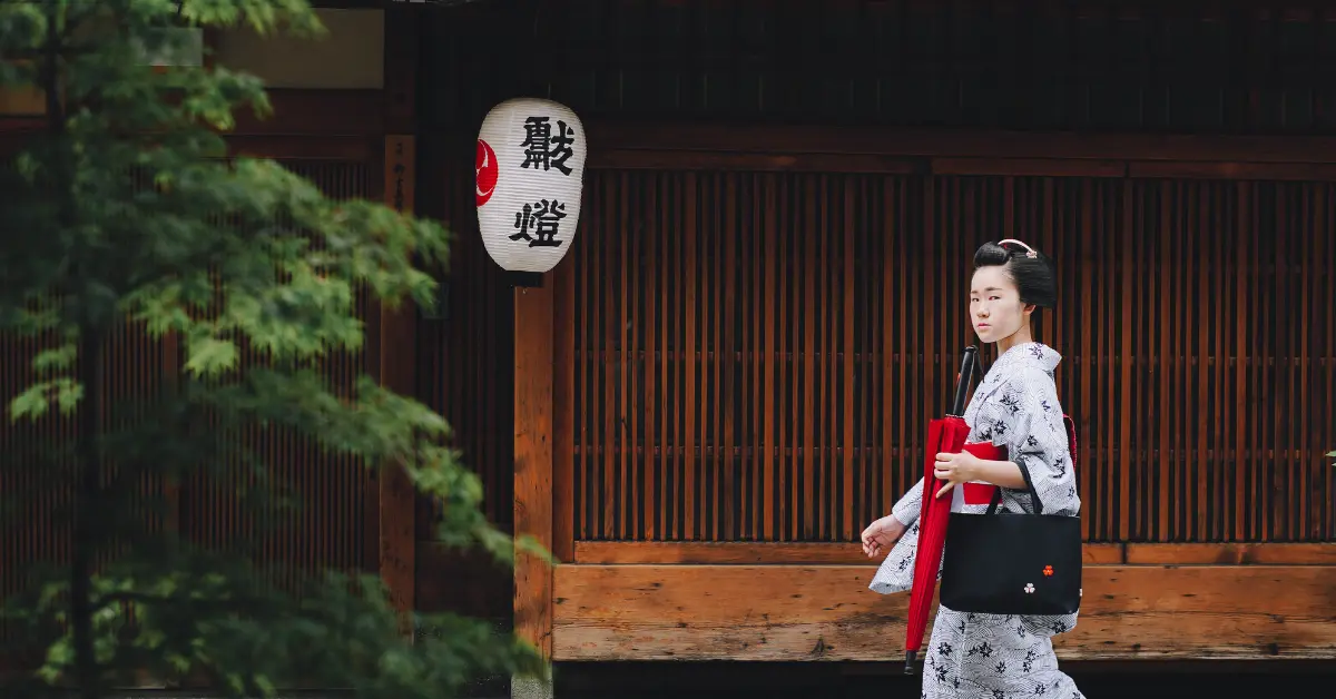 A woman in a traditional Japanese kimono walks past a wooden building with a paper lantern.