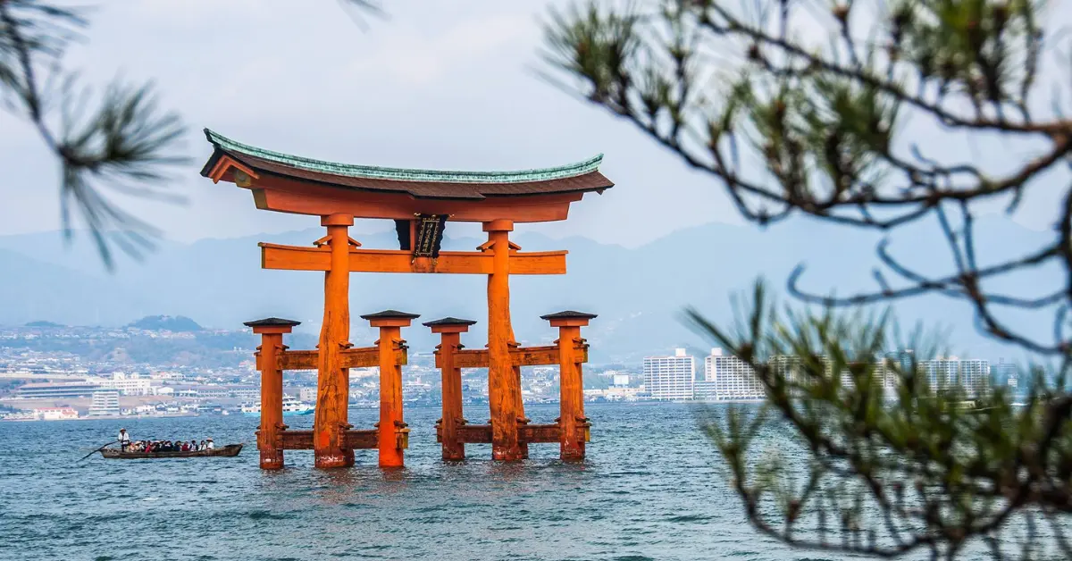 A large, iconic red torii gate stands in the water, framed by tree branches in the foreground. In the background, a boat with passengers floats on the water, and a cityscape with buildings and mountains is visible under a cloudy sky.