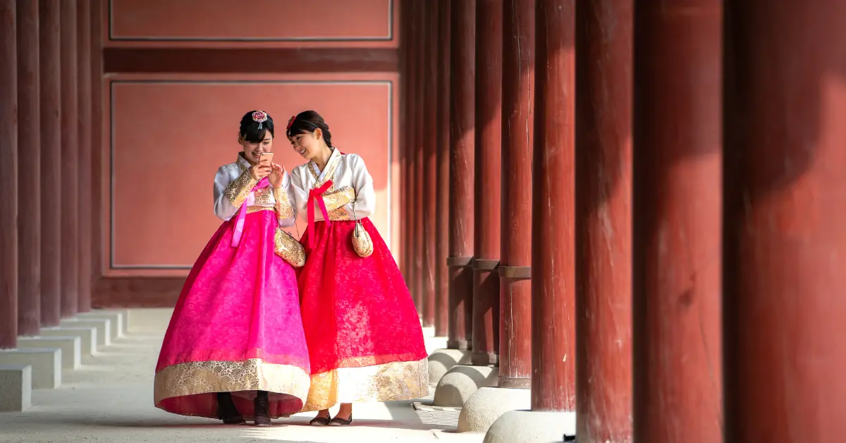 Two women dressed in traditional Korean hanbok stand in a corridor with red columns. They are smiling and looking at a smartphone together. The hanbok are vibrant pink with gold accents, and the corridor is bathed in soft, natural light.