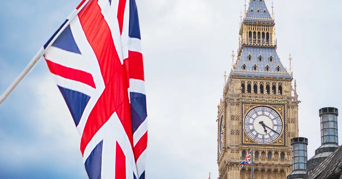 A Union Jack flag in the foreground with the Elizabeth Tower, commonly known as Big Ben, in the background. The sky is overcast, and the clock on the tower shows the time as 10:10. The image captures iconic symbols of the United Kingdom.