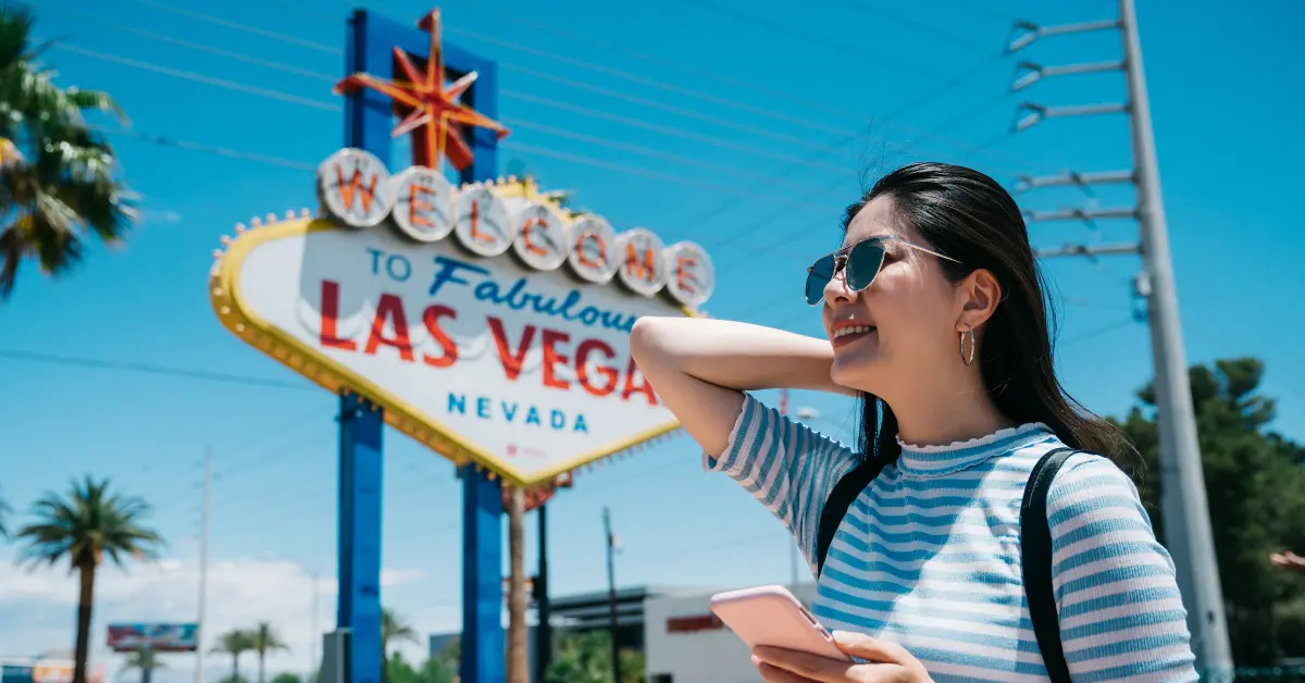 A person smiling and standing in front of the "Welcome to Fabulous Las Vegas, Nevada" sign. They are wearing sunglasses, a striped shirt, and a backpack, holding a smartphone. The sky is clear and blue, with palm trees and power lines in the background, indicating a sunny day.