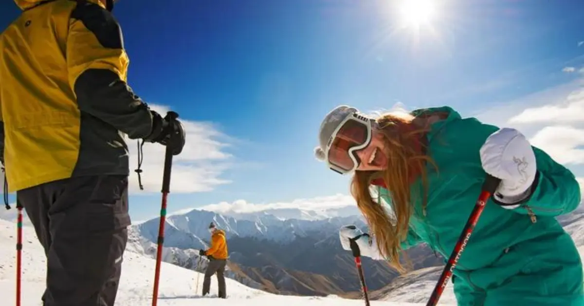 Three people enjoying a sunny day skiing on a snowy mountain in New Zealand, with one person in the foreground smiling and leaning towards the camera, showcasing the fun and excitement of the adventure.