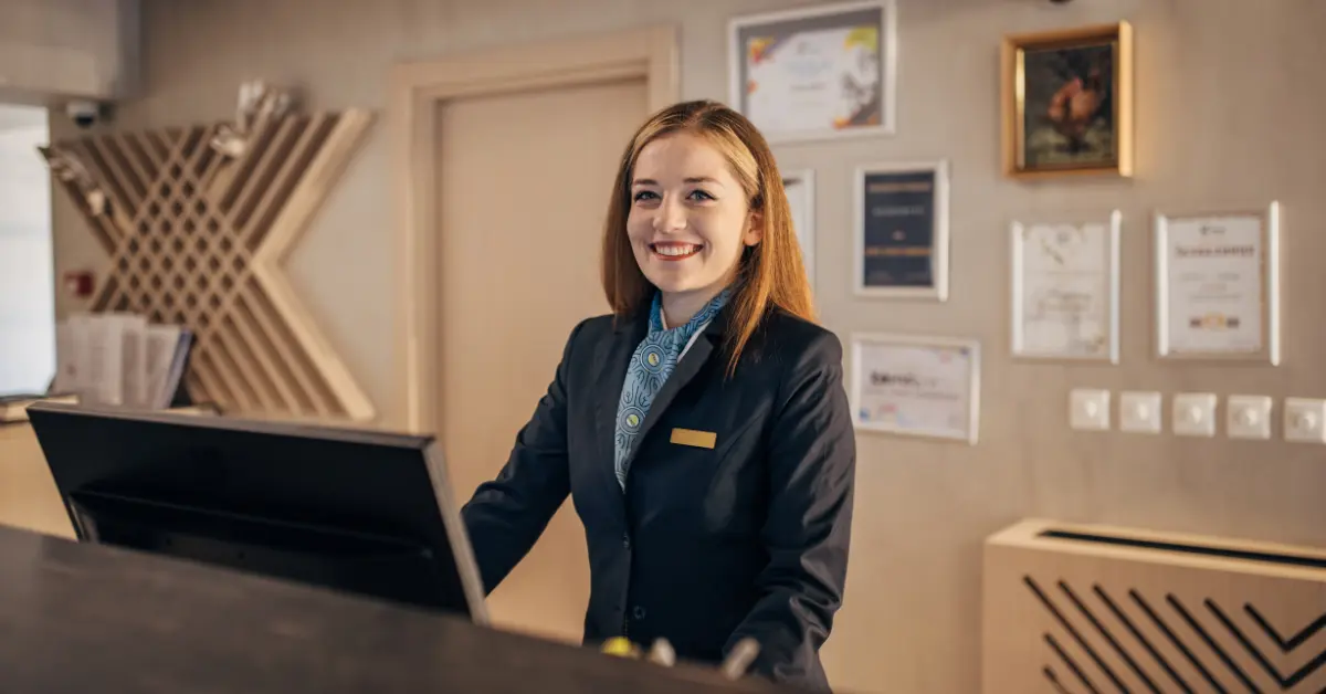 Smiling receptionist in a professional uniform standing behind a modern reception desk, with framed certificates and awards on the wall, representing a working holiday in the USA.