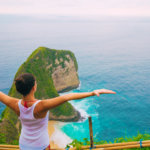 Woman standing on a clifftop with arms outstretched, enjoying a breathtaking view of a turquoise ocean and rocky islands.
