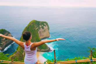 Woman standing on a clifftop with arms outstretched, enjoying a breathtaking view of a turquoise ocean and rocky islands.