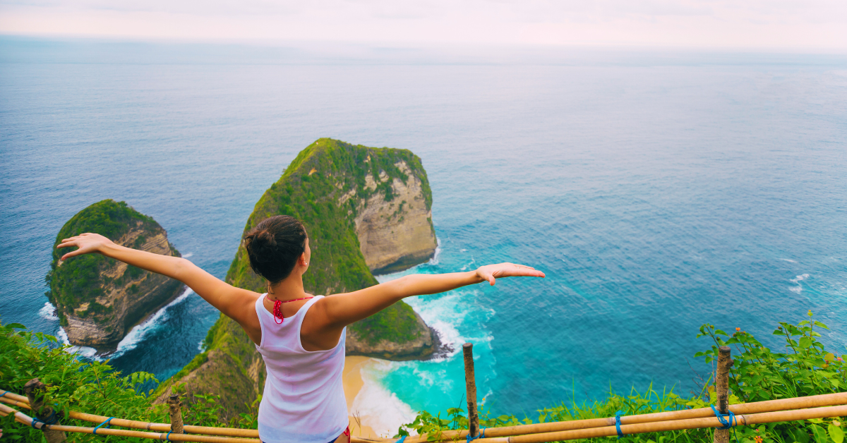 Woman standing on a clifftop with arms outstretched, enjoying a breathtaking view of a turquoise ocean and rocky islands.